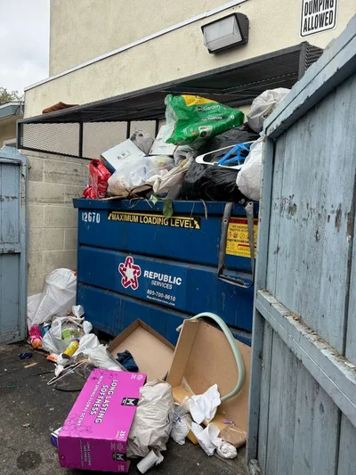 Overflowing blue dumpster with mixed household junk, cardboard boxes, and waste materials cleared from residential property
